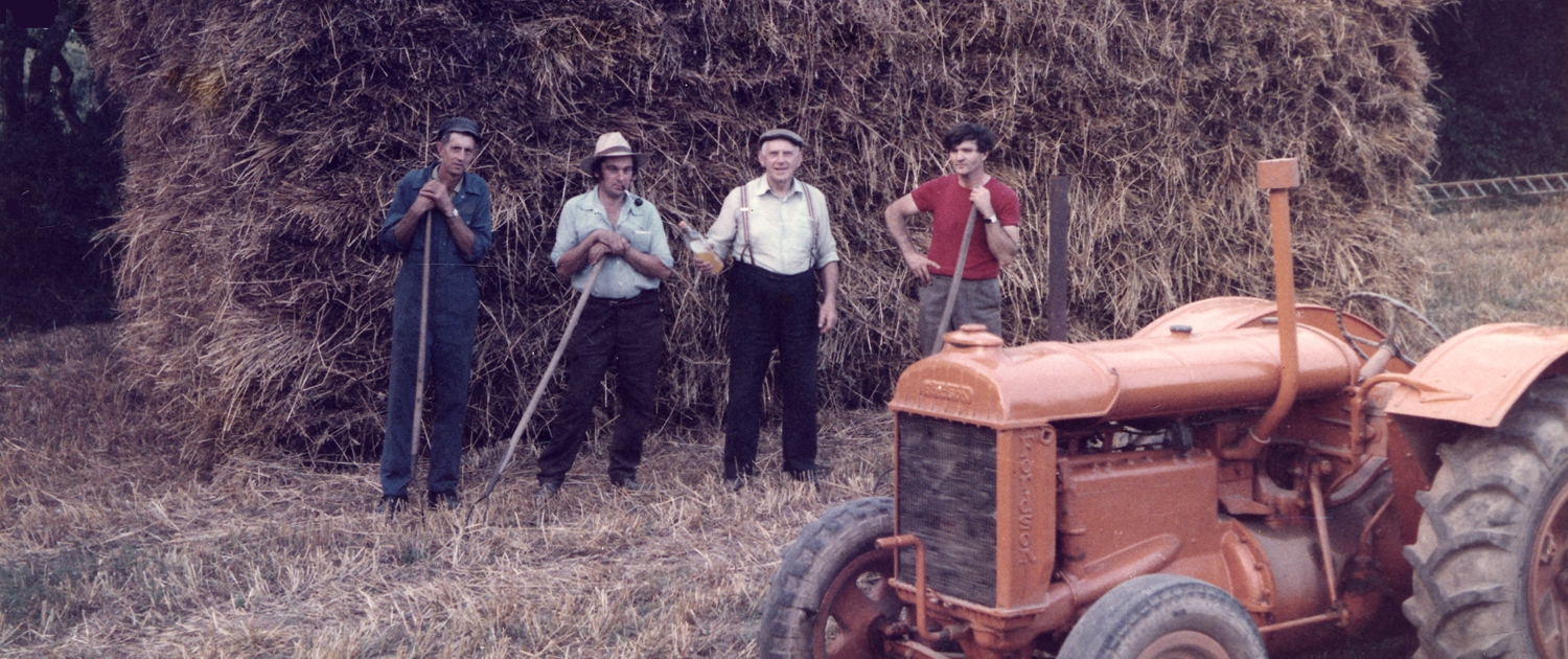 Farm workers in front of a hayrick, resting with their tools, orange tractor in the foreground